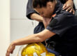 Side view of late 30s woman in uniform sitting on bench in fire station locker room with helmet in lap, head down, and eyes closed.